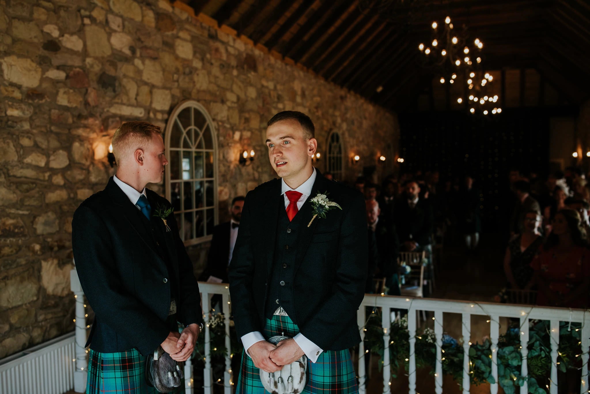 Two grooms in traditional green tartan kilts and black jackets stand at the front of a rustic stone wedding venue during a ceremony.