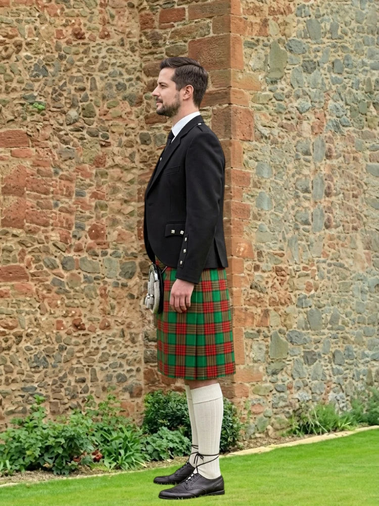 Man wearing a traditional Scottish kilt standing against a stone wall.