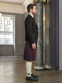 Man wearing a black jacket and red plaid kilt in an elegant room with marble columns.