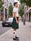 A man standing on a city sidewalk wearing a white t-shirt and a dark green pleated kilt, paired with white socks and black leather shoes. A black Mercedes-Benz car and a bicycle are parked in the background of the street scene.