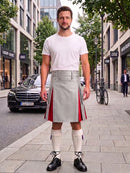 A man stands on a city sidewalk wearing a white t-shirt, a light gray kilt with red side panels, white knee-high socks, and black shoes. Buildings, trees, a parked black car, and a bicycle are visible in the background.