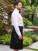 A man wearing a black kilt with silver hardware, a white dress shirt, and a black bow tie, standing in a landscaped garden with plants and a water feature in the background.