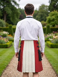 A man viewed from behind wearing a white long-sleeve shirt and a pleated kilt with vertical panels in red, off-white, and black, standing on a garden path.