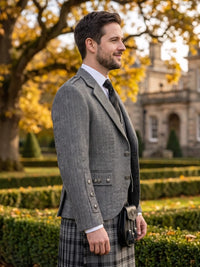 A man in profile wearing a gray herringbone blazer, white dress shirt, dark tie, and a gray plaid kilt with a sporran, standing outdoors in front of a stone building and autumn trees.