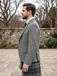 A man in profile wearing a gray herringbone blazer and a black-and-gray plaid kilt, standing outdoors on a stone patio with bare trees and a stone wall in the background.