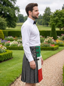 A man standing in a garden wearing a gray pleated kilt with a green, white, and red vertical panel on the back, a white dress shirt, and a black bow tie.