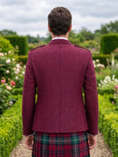 A man viewed from behind wearing a textured burgundy kilt jacket and a red, green, and black tartan kilt, standing in a garden path.