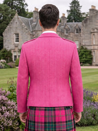 A man viewed from behind wearing a bright pink textured herringbone blazer and a pink, green, and black plaid kilt, standing in front of a large stone castle on a green lawn.