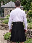 A man viewed from behind wearing a black pleated kilt and a white long-sleeve dress shirt, standing in a garden with stone walls and a house in the background.