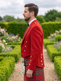 A man in profile wearing a bright red formal jacket with silver buttons and a red tartan kilt, standing on a garden path with trimmed hedges and flowers in the background.