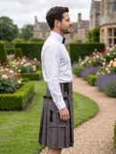 A man in profile wearing a white dress shirt and a dark gray pleated kilt with black straps, standing in a landscaped garden path with hedges and flowers in the background.