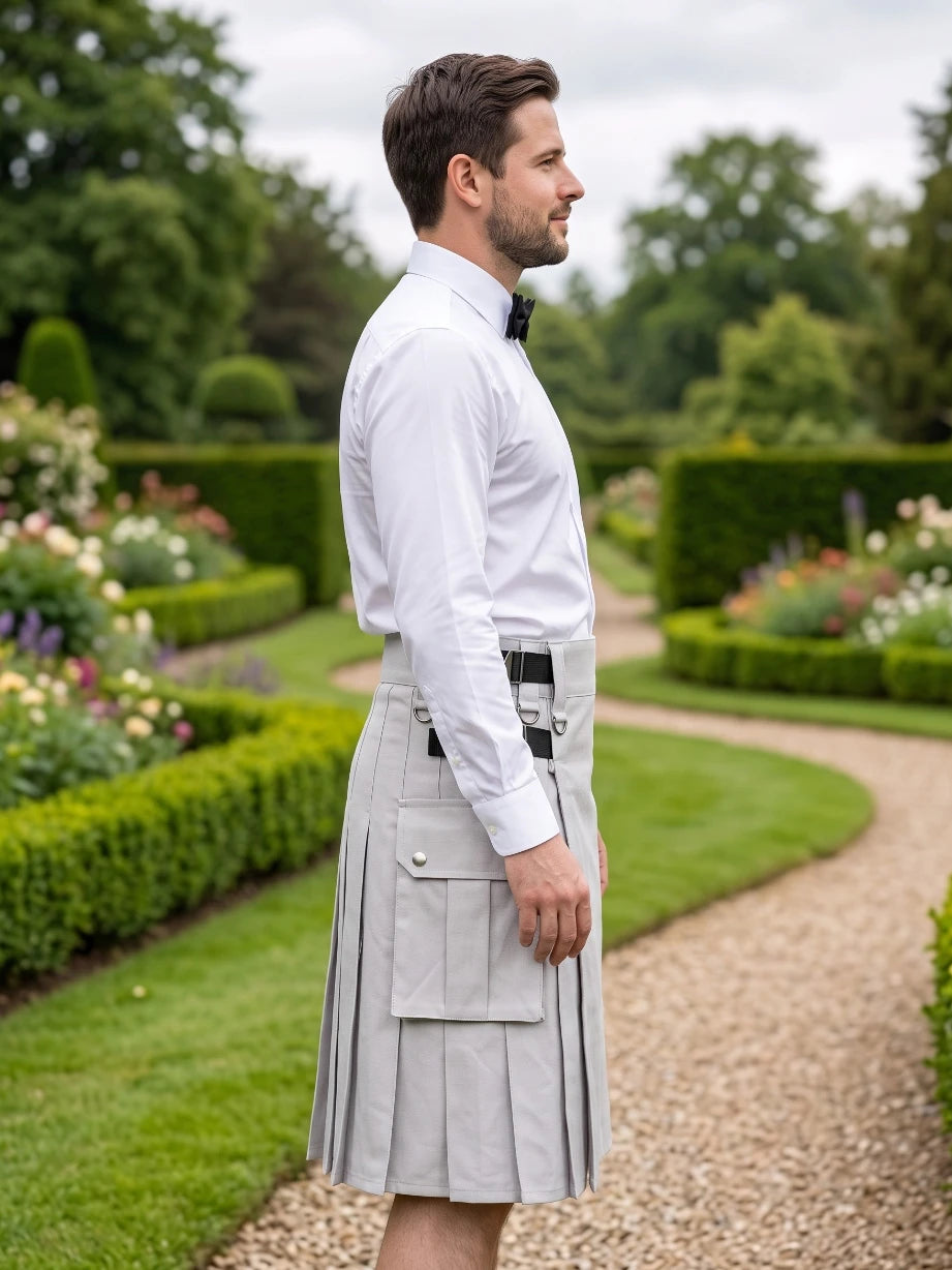 A man in a white dress shirt and black bow tie stands in profile outdoors wearing a light gray pleated kilt with flap pockets and a black belt, set against a manicured garden path and hedges.
