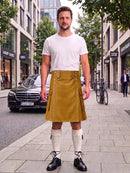 A man stands on a city sidewalk wearing a white crew-neck t-shirt and a mustard yellow corduroy kilt with silver metal accents, paired with white knee-high socks and black leather shoes.