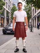 A man stands on a city sidewalk wearing a brown leather kilt, a white t-shirt, white knee-high socks, and black leather shoes. The background shows a blurred street scene with buildings, trees, a parked black car, and a bicycle.
