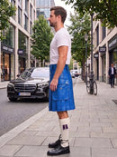 A man standing on a city sidewalk wearing a bright blue leather kilt, a white t-shirt, white socks, and black shoes. The background shows a street with cars, trees, and buildings.