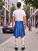 A man standing on a city sidewalk, viewed from behind, wearing a white t-shirt, a bright blue pleated leather kilt, white knee-high socks, and black shoes. Buildings, trees, a black Mercedes, and a bicycle are visible in the background.