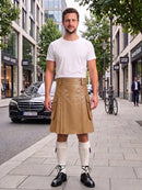 A man stands on a city sidewalk wearing a tan pleated leather kilt with silver-tone hardware, a white crew-neck t-shirt, white knee-high socks, and black leather oxford shoes. The background shows a street scene with a black Mercedes-Benz, storefronts, and trees.