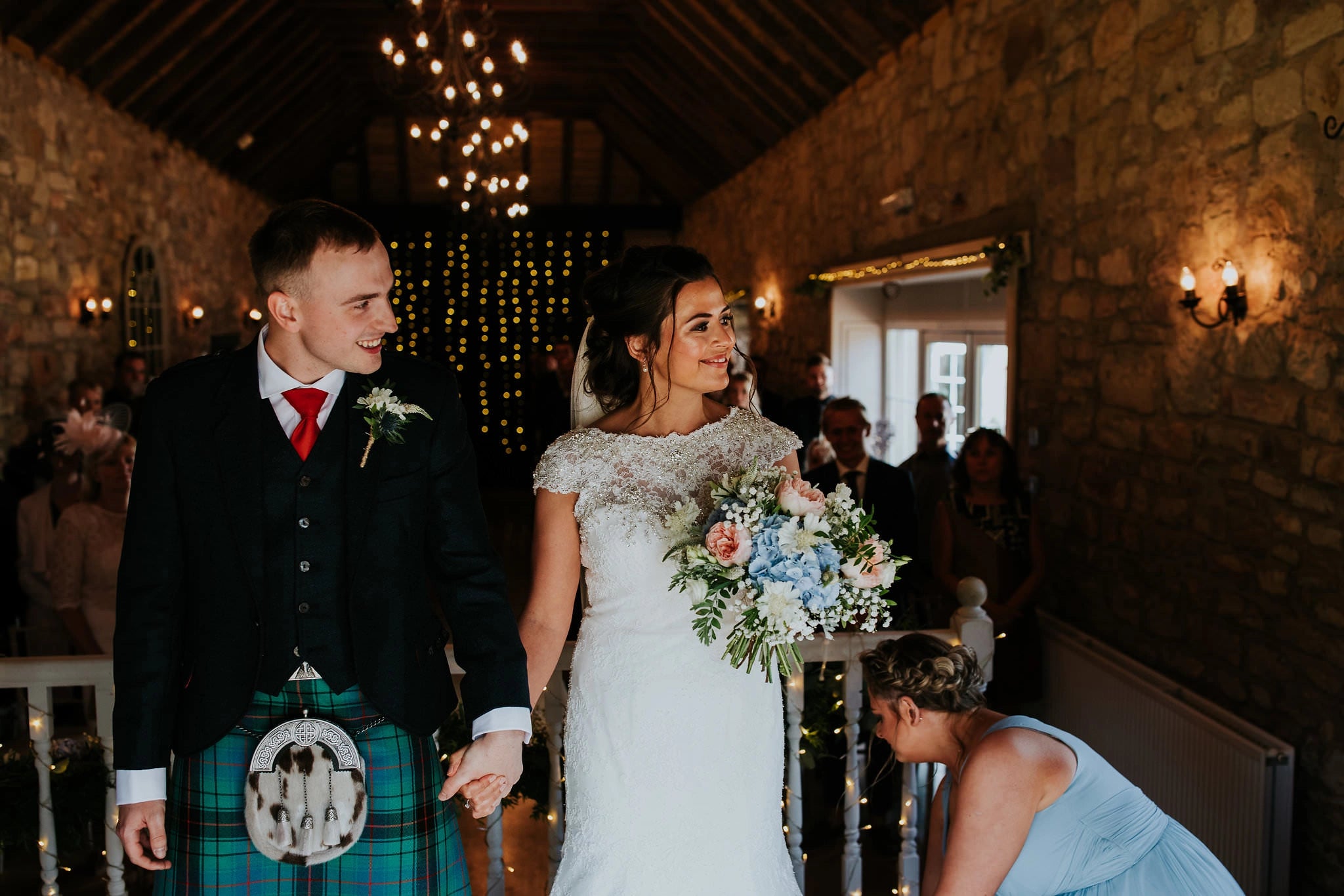 Bride and groom walking hand in hand down the aisle after a wedding ceremony in a rustic barn venue, with warm lights and floral bouquet