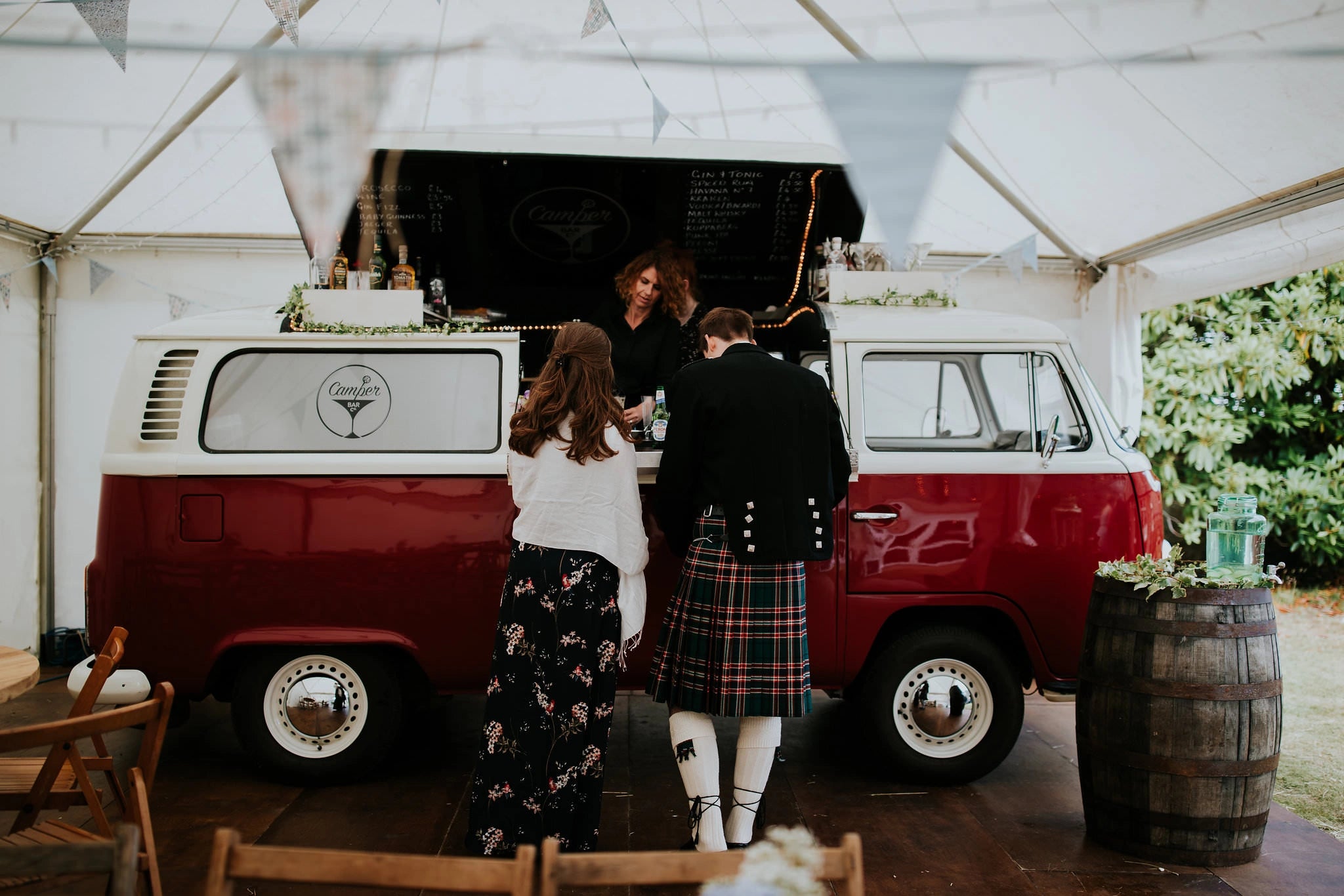 Couple at a vintage red VW campervan bar, with the groom wearing a traditional tartan kilt at a rustic wedding reception.